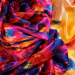 Fular de seda natural XL de 110x180 cm con estampado artístico en tonos amarillos, rojos y azules, anudado al hombro sobre conjunto azul de Julunggul